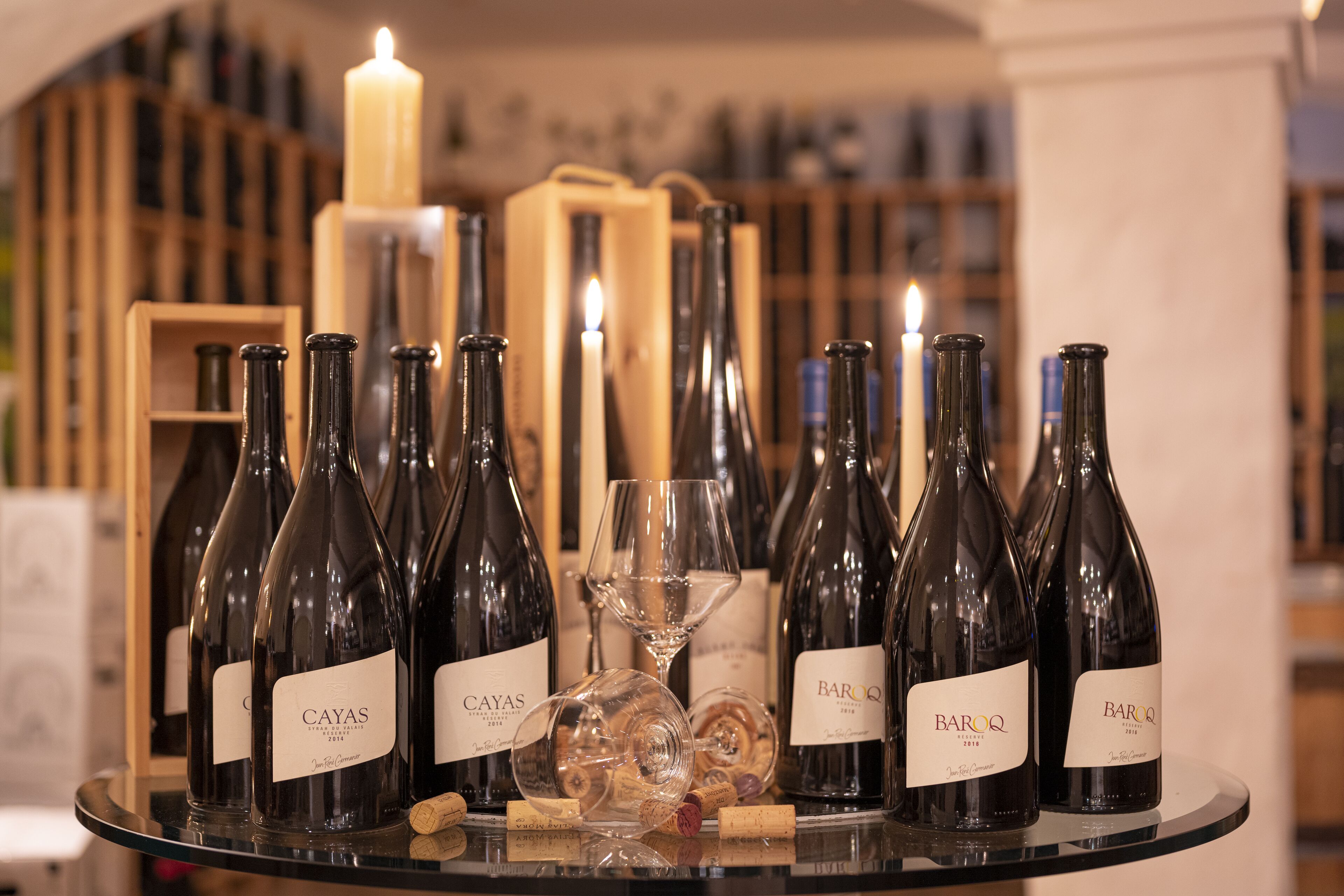 A selection of wine bottles on a glass table with corks, surrounded by wine glasses and a lit candle, set against a backdrop of shelves with bottles.