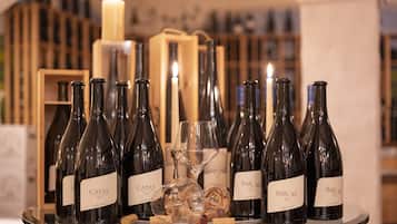 A selection of wine bottles on a glass table with corks, surrounded by wine glasses and a lit candle, set against a backdrop of shelves with bottles.