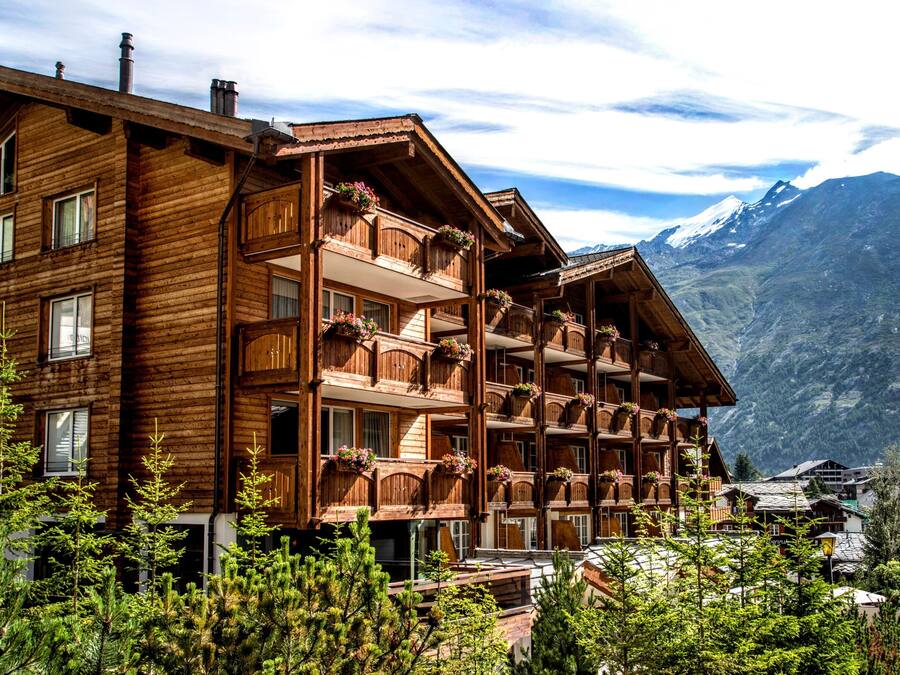 A multi-story wooden building with balconies, surrounded by trees and mountains in the background.