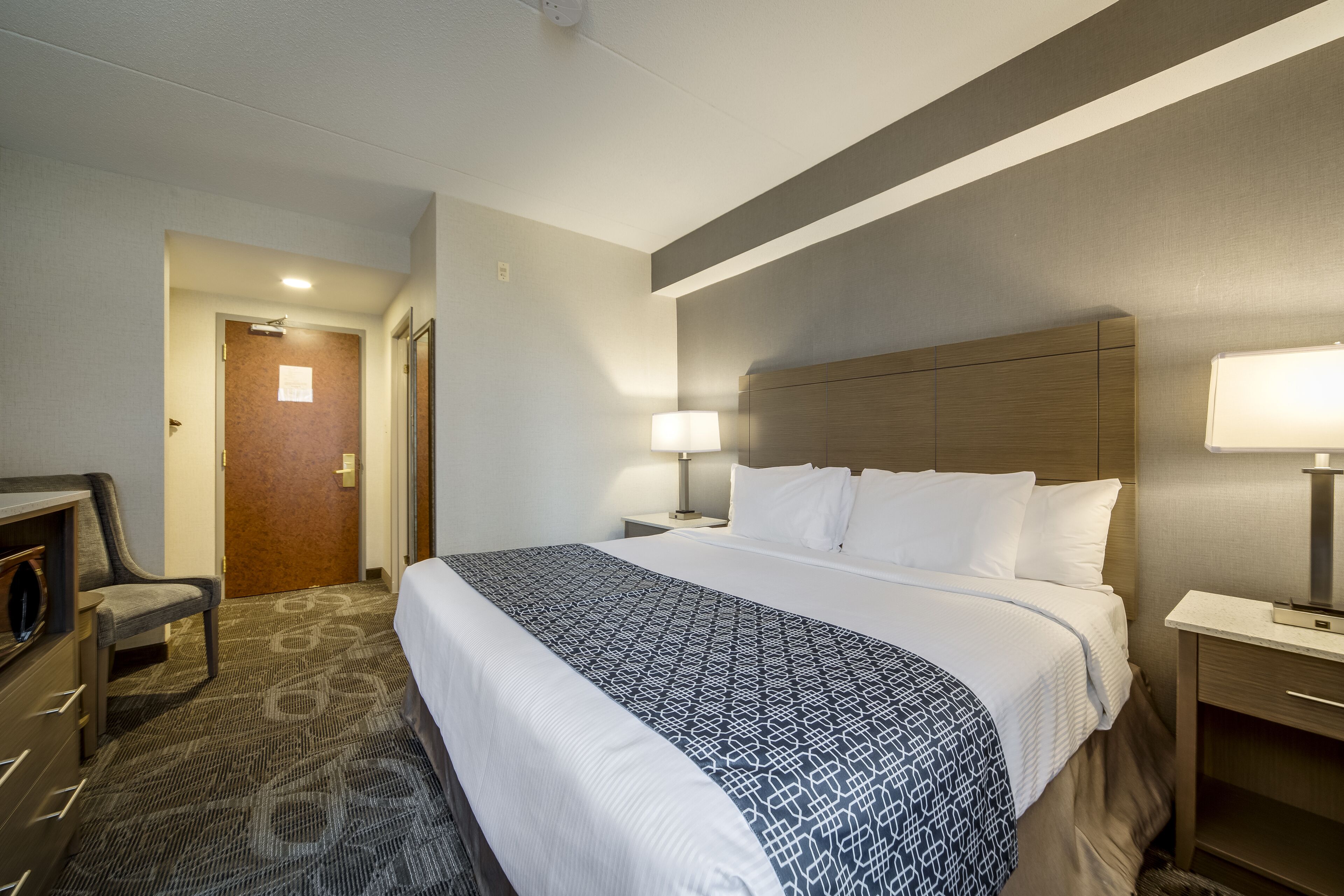 A hotel room with a large bed, two bedside tables with lamps, a chair, and a carpeted floor.