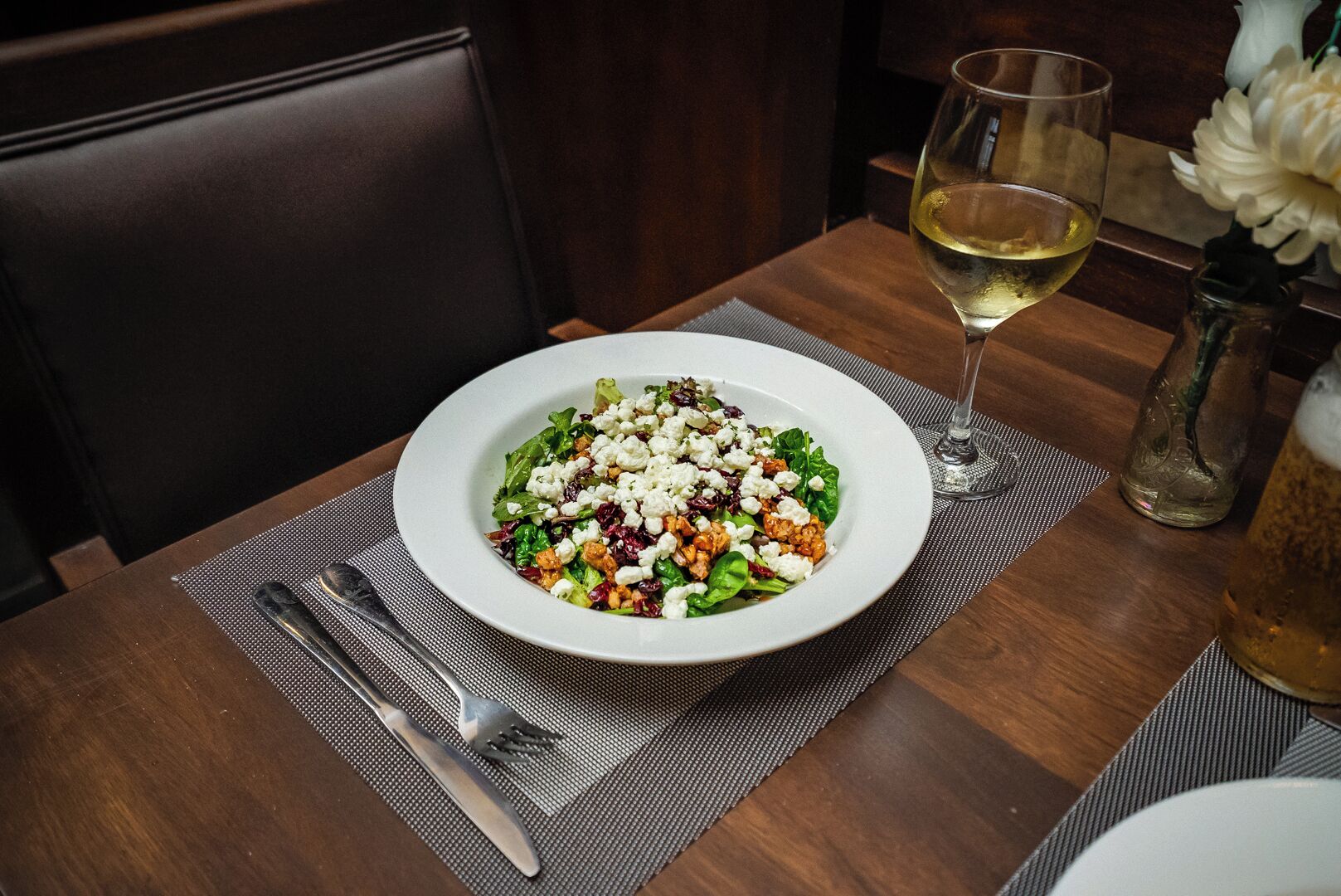 Una mesa de comedor con un plato blanco de ensalada, una copa de vino blanco y un jarrón con flores blancas.