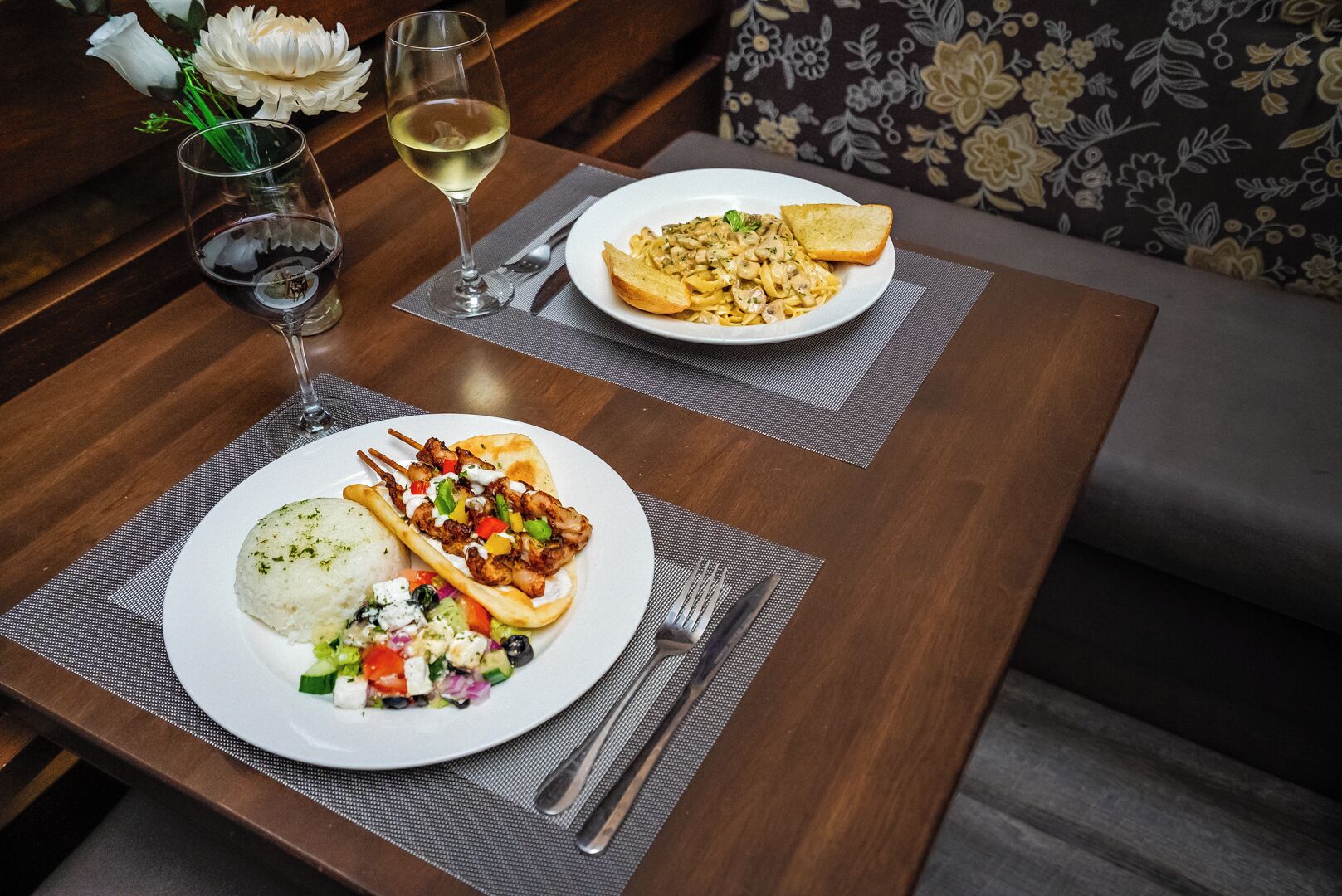 A dining table with two plates of food, a glass of white wine, and a vase with white flowers.