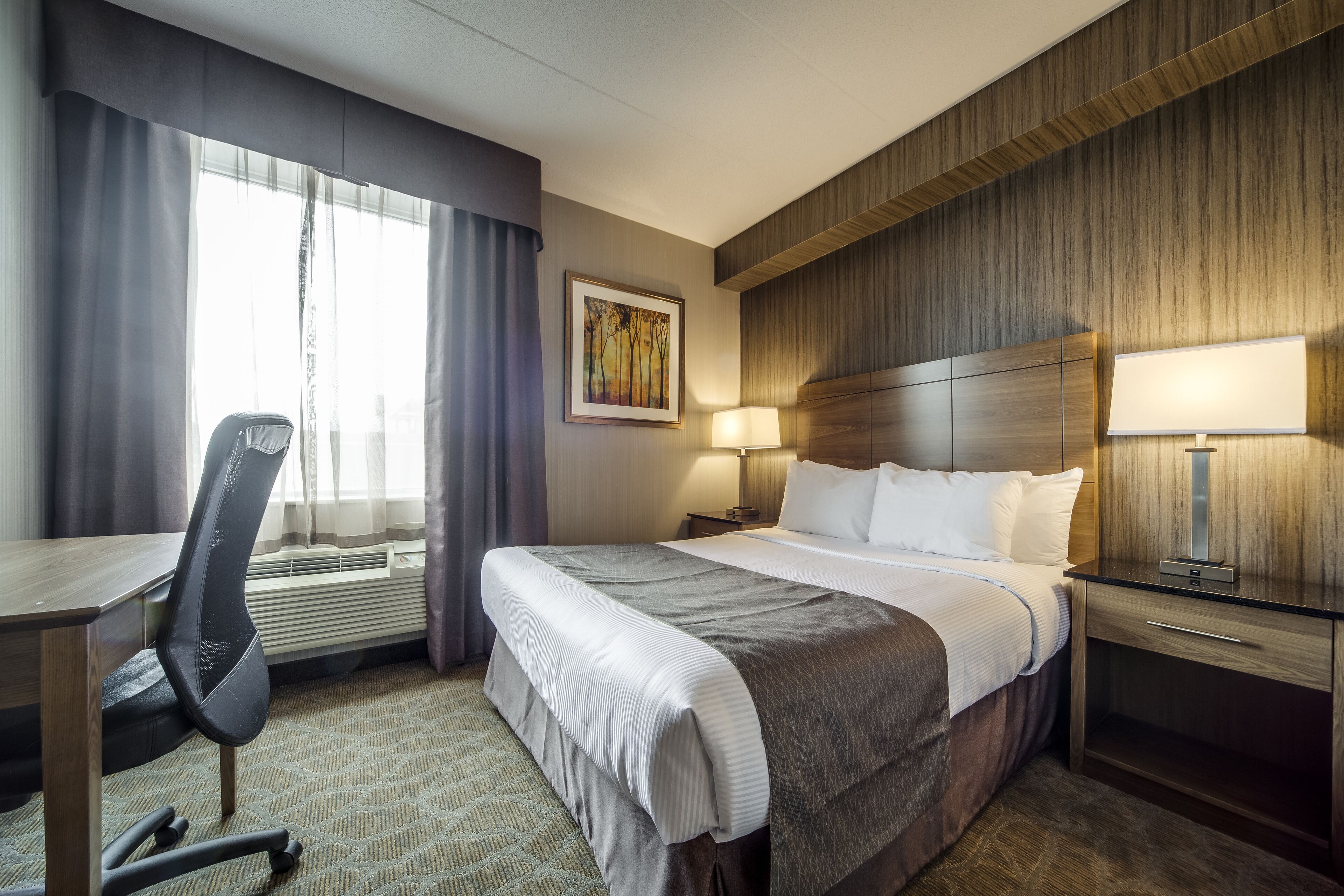 A hotel room with a large bed, a desk, and a chair. There is a window with curtains, a painting on the wall, and a lamp on the nightstand.