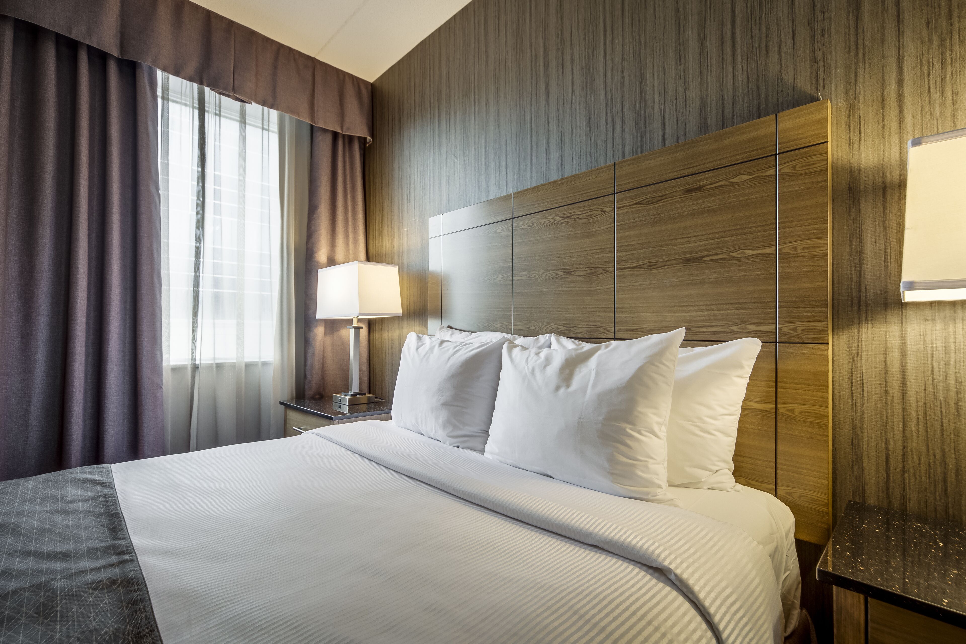 Ein Hotelzimmer mit einem großen Bett, einem Holzkopfstück, einem Nachttisch mit Lampe und einem Fenster mit Vorhängen.