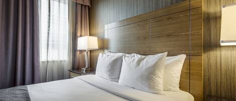 A hotel room with a large bed, a wooden headboard, a nightstand with a lamp, and a window with curtains.
