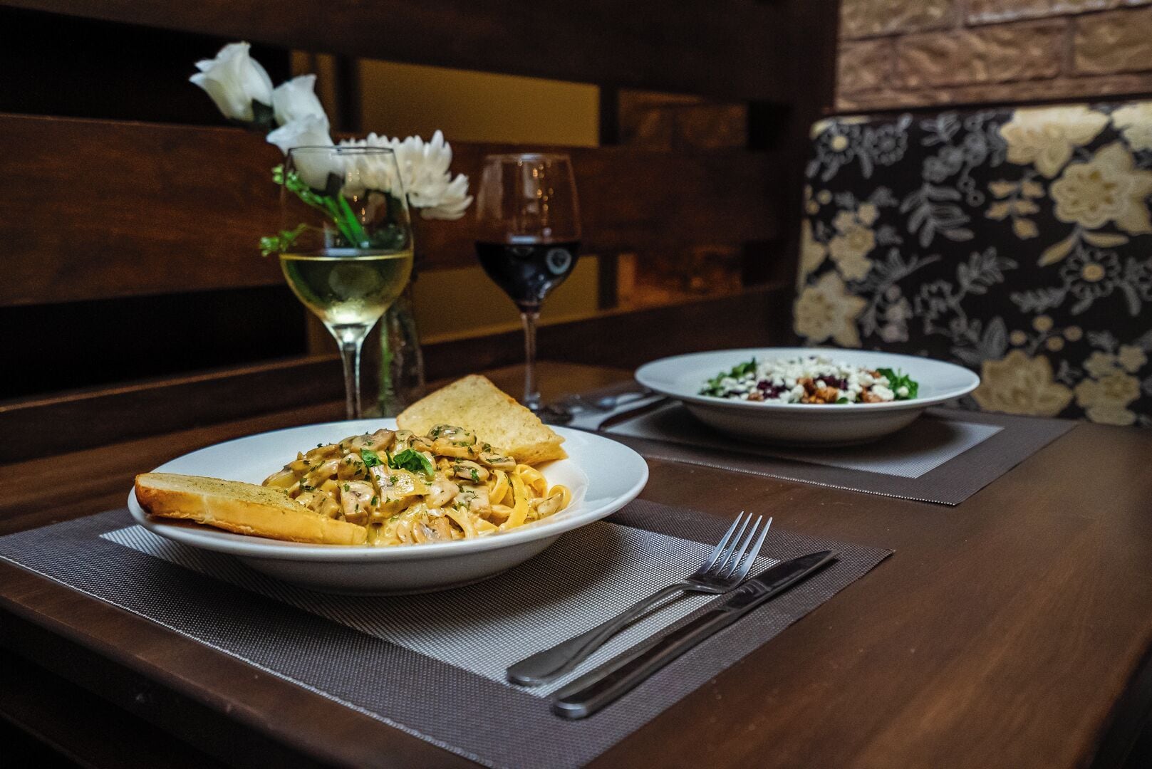 Une table à manger avec une assiette de pâtes et un saladier rempli de salade, un verre de vin et un vase avec des fleurs.