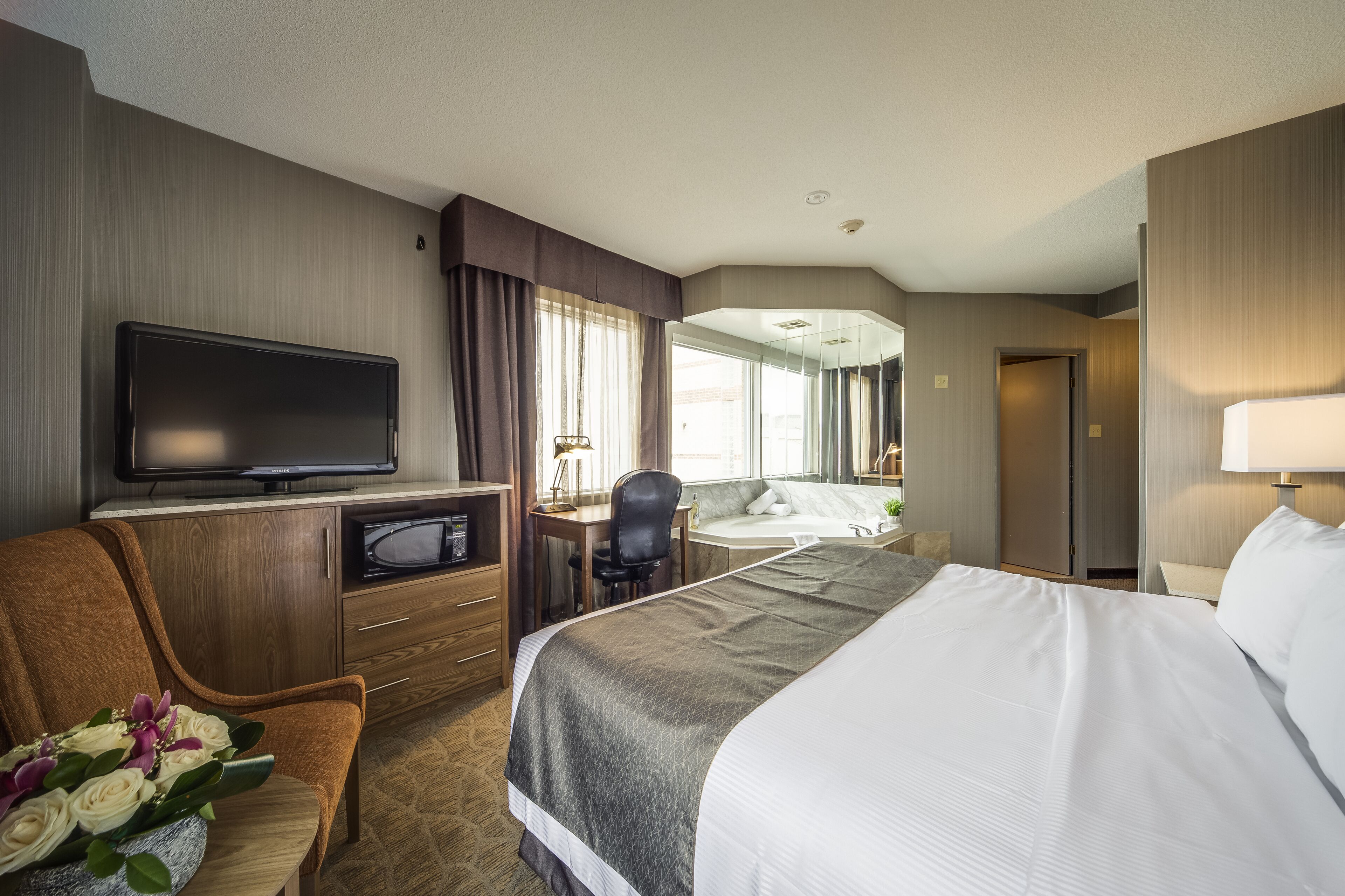 A hotel room with a large bed, a desk with a computer, a television, and a seating area with a chair and a bouquet of flowers.