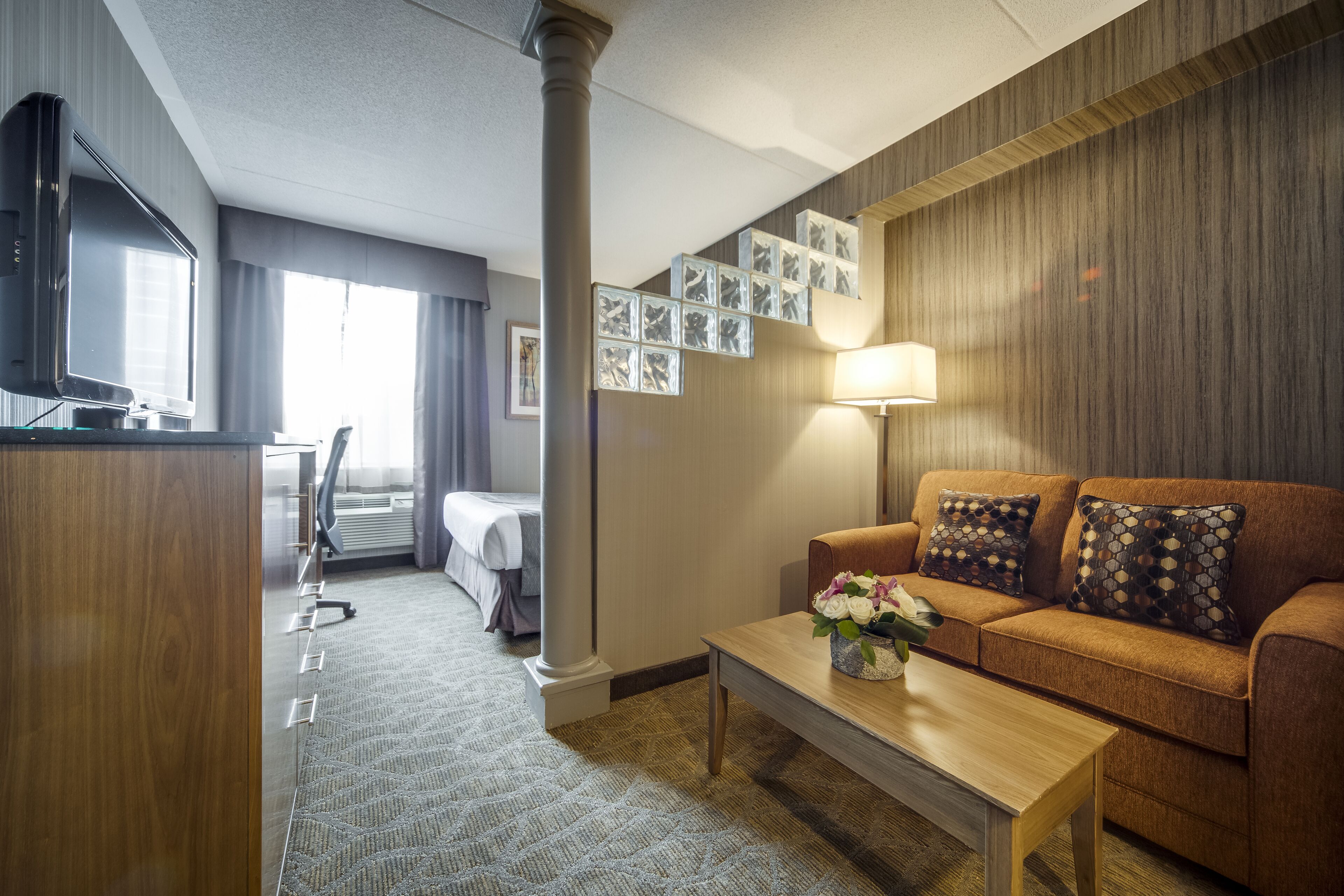 Habitación de hotel con sofá de madera, una mesa de centro, televisor, escritorio con silla y una ventana grande con cortinas.