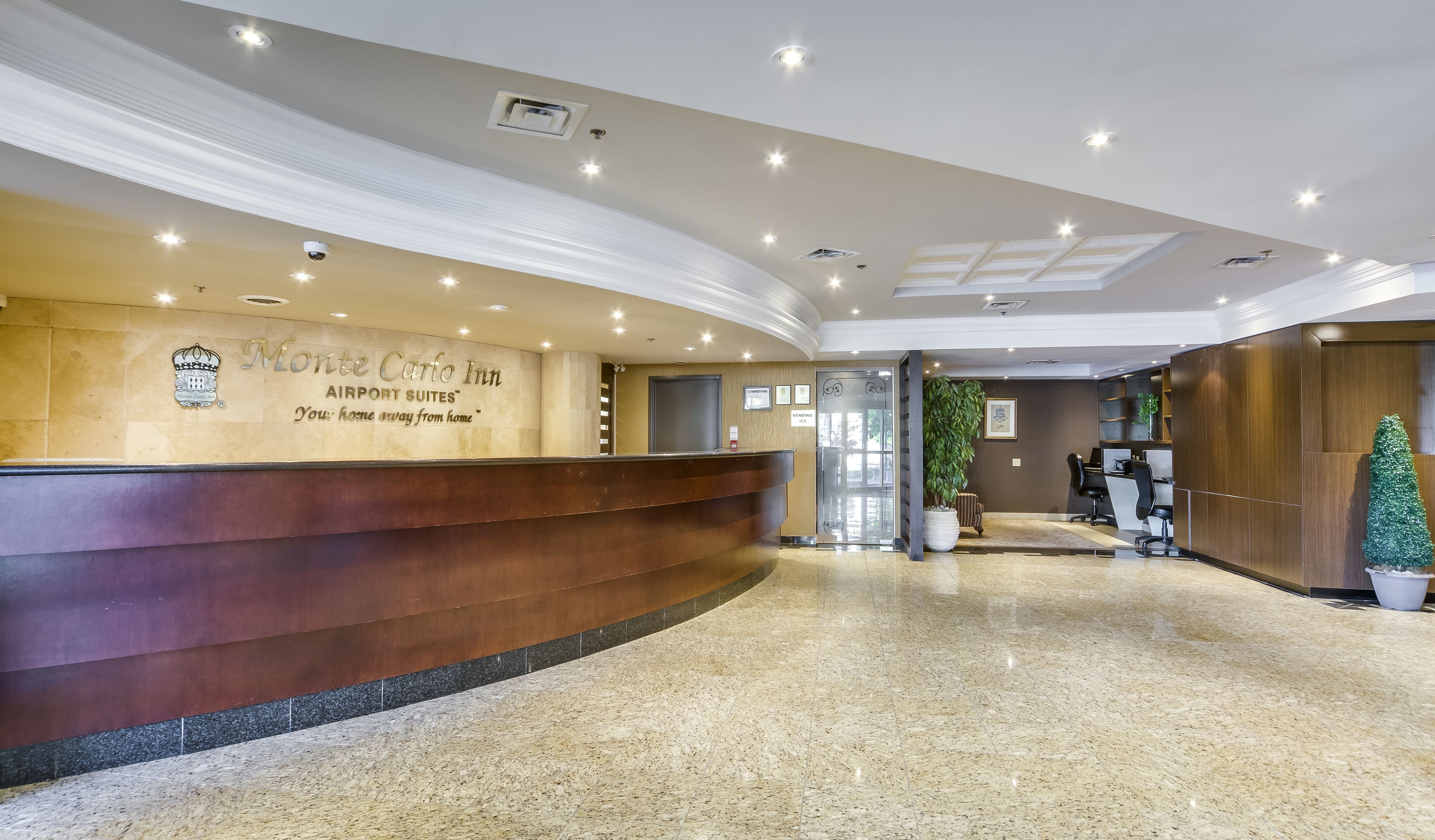 Zona de recepción de hotel con un mostrador curvo, un letrero en la pared para Monte Carlo Inn Airport Suites, una máquina expendedora y una planta en maceta.