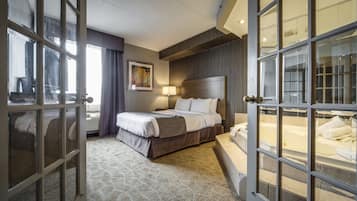 Habitación de hotel con una cama grande, cabecera, mesita de noche, lámpara y vistas a una bañera.
