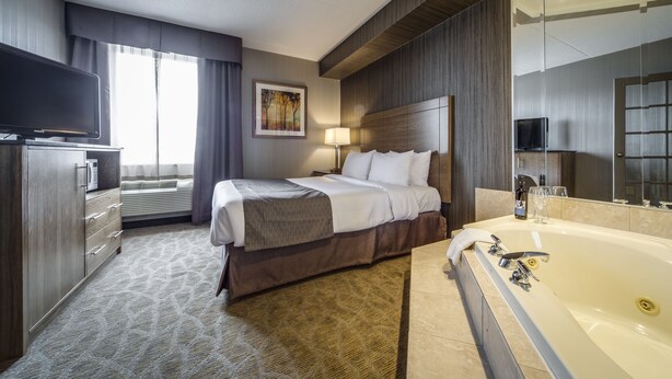 A hotel room with a large bed, a TV, a desk with a computer, a large window, and a bathroom with a bathtub and mirror. - Monte Carlo Inn Airport Suites (Mississauga)
