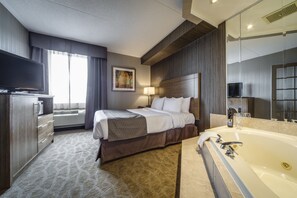 A hotel room with a large bed, a TV, a desk with a computer, a large window, and a bathroom with a bathtub and mirror. - Monte Carlo Inn Airport Suites (Mississauga)