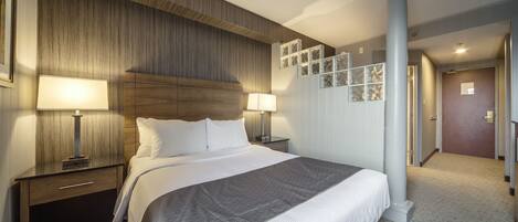 A hotel room with a large bed, two bedside tables with lamps, a wooden headboard, and a hallway with a column and framed pictures on the wall.