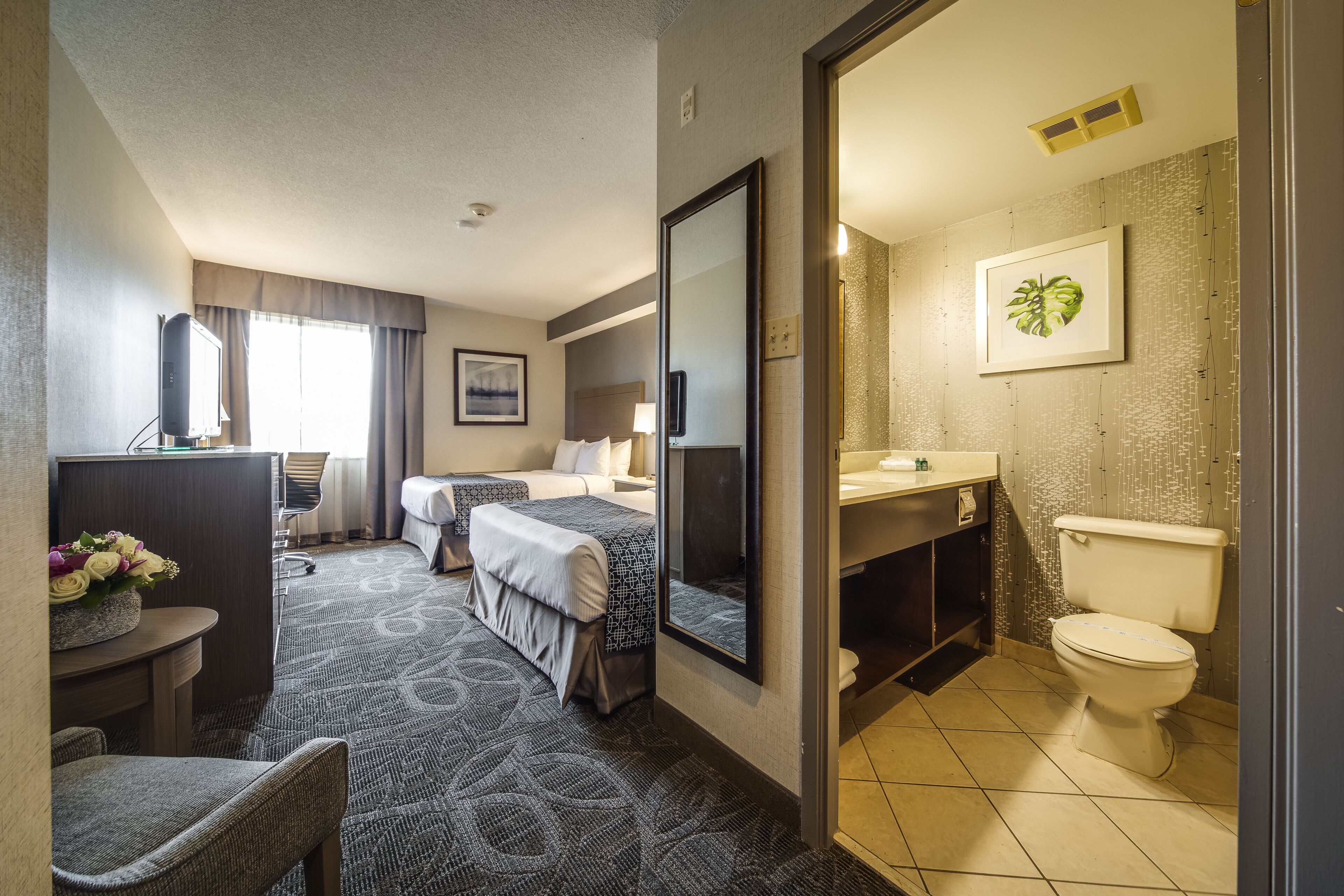 A hotel room with a bed, a desk, a chair, a bathroom with a toilet and sink, and a large mirror.