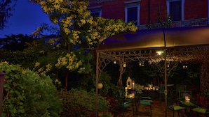 A covered outdoor seating area with tables and chairs, surrounded by lush greenery and a red brick building in the background. - Hotel Bagliori (Milan)