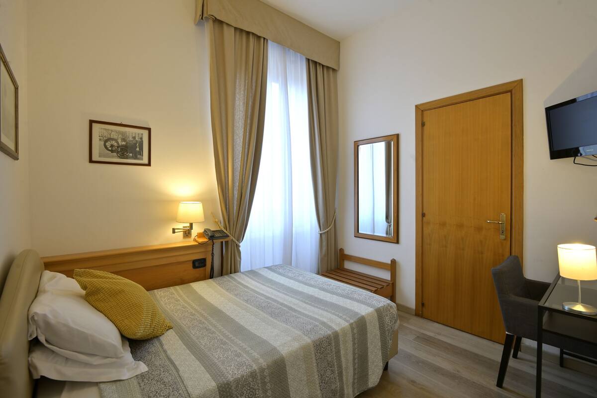 A hotel room with a bed, bedside table, lamp, chair, wooden door, and a television mounted on the wall.