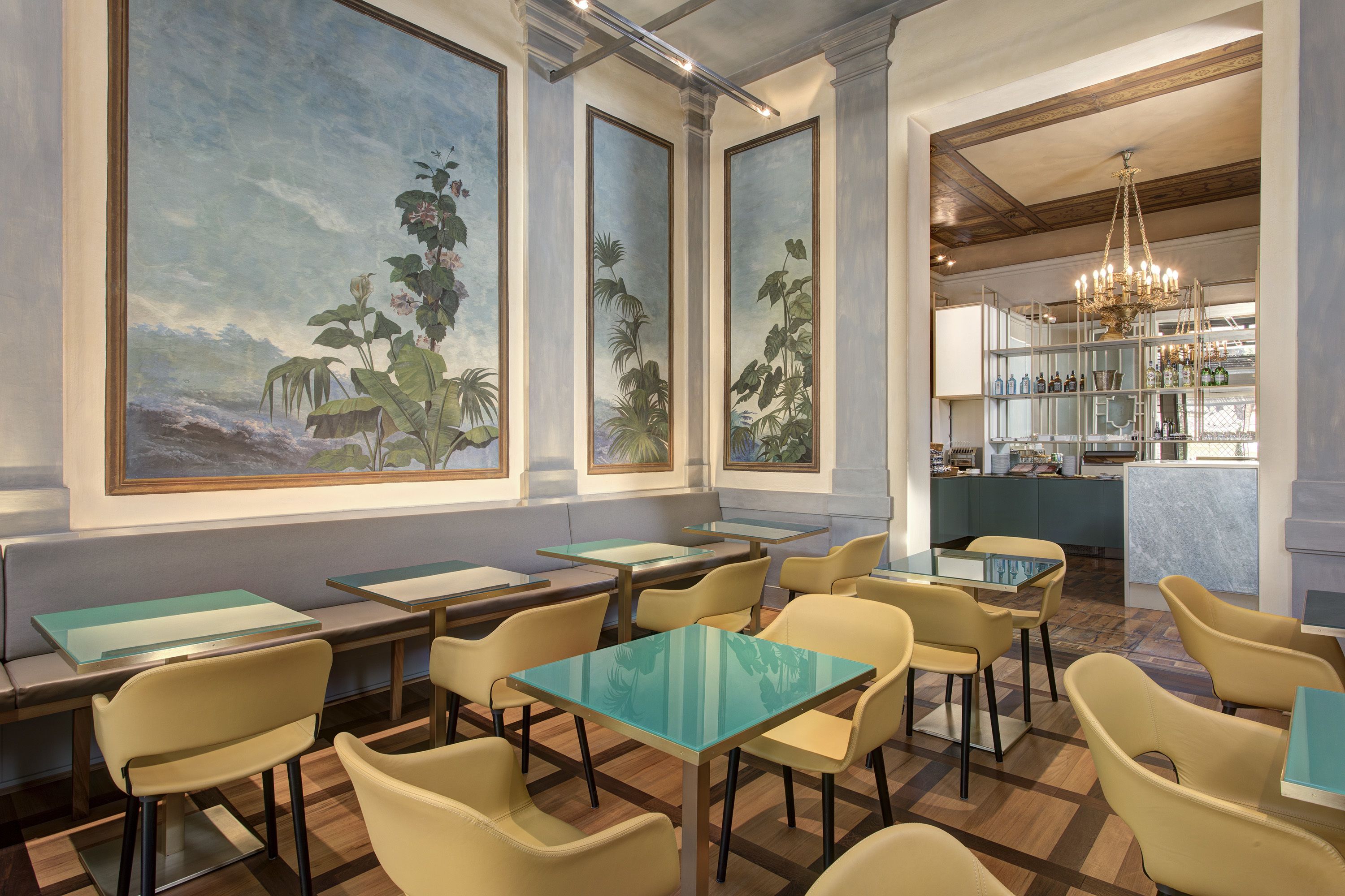 A restaurant with high ceilings, large windows with tropical artwork, and a bar area with shelves of bottles.