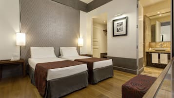 A hotel room with two beds, a small seating area, a bathroom with a mirror and sink, and a hallway leading to other rooms.