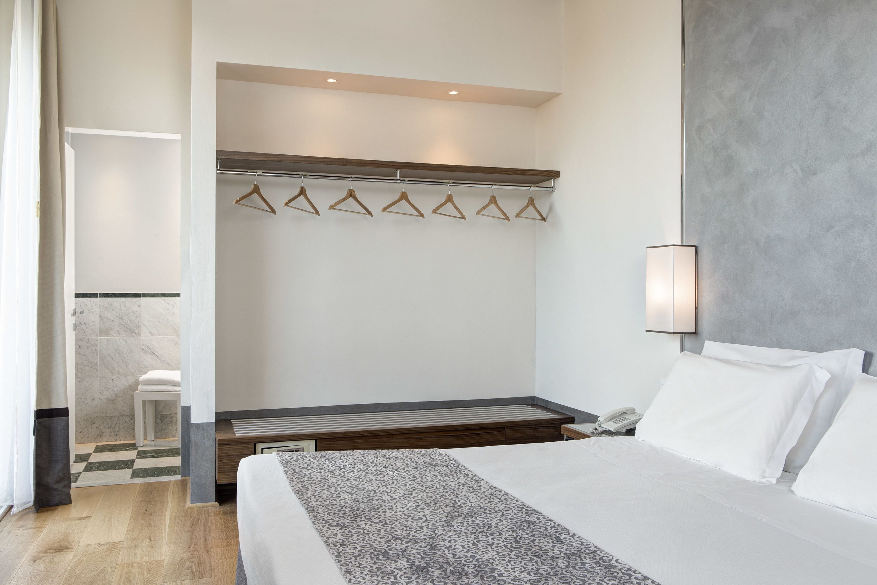 Ein Hotelzimmer mit einem Bett, einem Nachttisch, einem Telefon, einem Kleiderständer mit Kleiderbügeln sowie einem Badezimmer mit Toilette und Waschbecken.