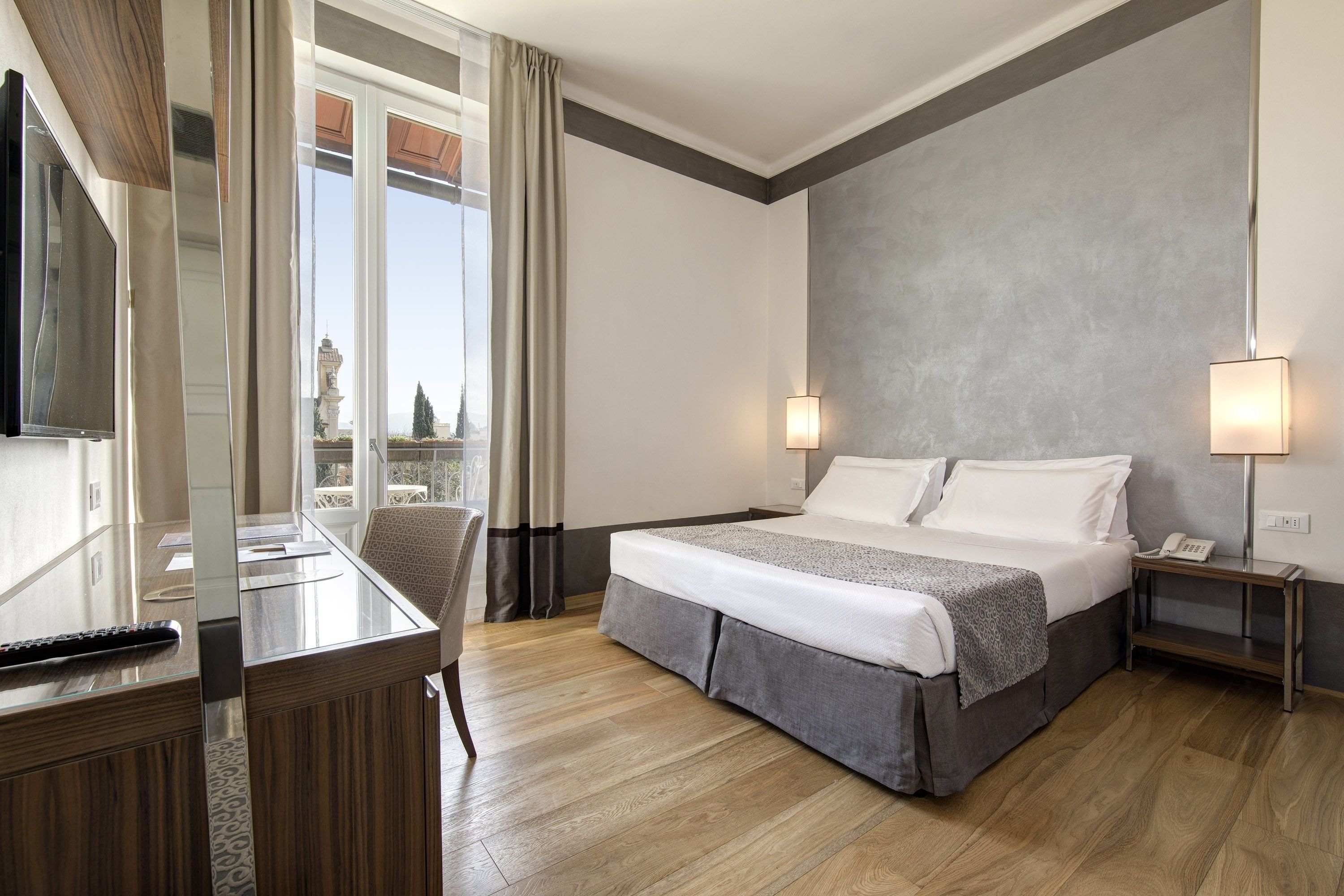 A hotel room with a bed, bedside tables, a desk, and a large window with curtains.