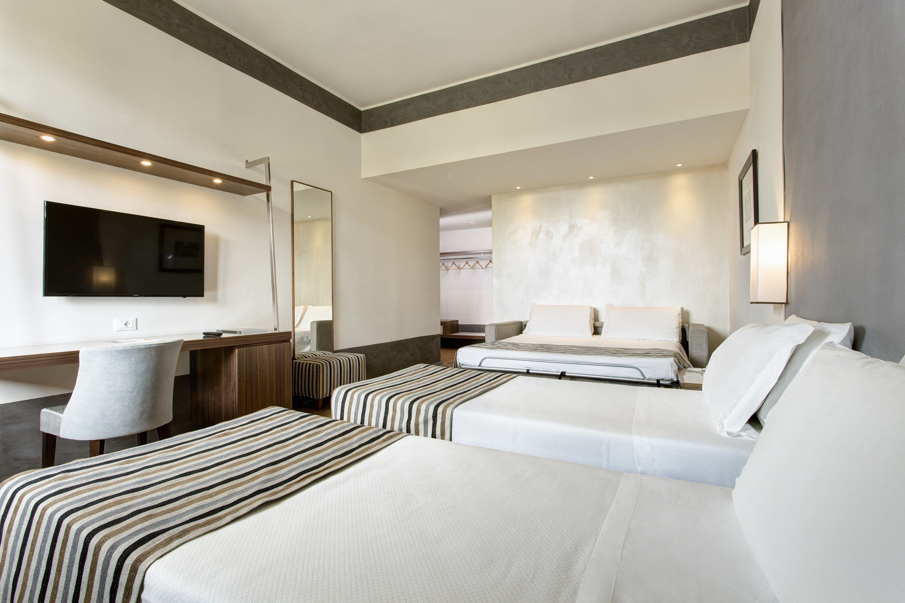 Ein Hotelzimmer mit zwei Betten, einem Schreibtisch, einem Sessel, einem Fernseher und einem großen Spiegel.