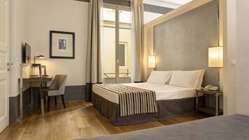 Een hotelkamer met een bed, nachtkastje met een lamp, een bureau met stoel, en een lijst met afbeelding aan de muur.