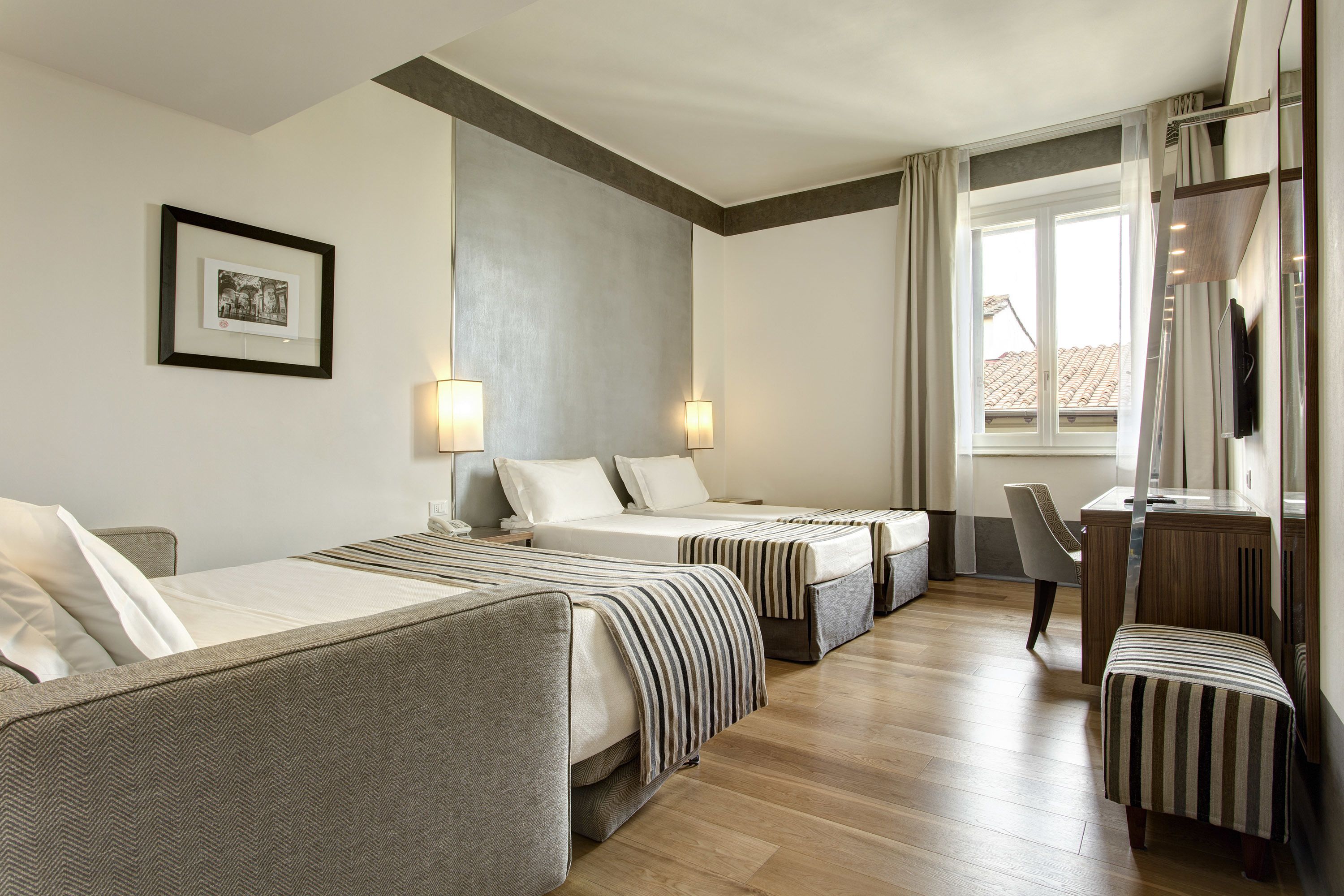 A hotel room with three beds, a desk, and a chair. There is a window with curtains, a picture on the wall, and a lamp on each bed.
