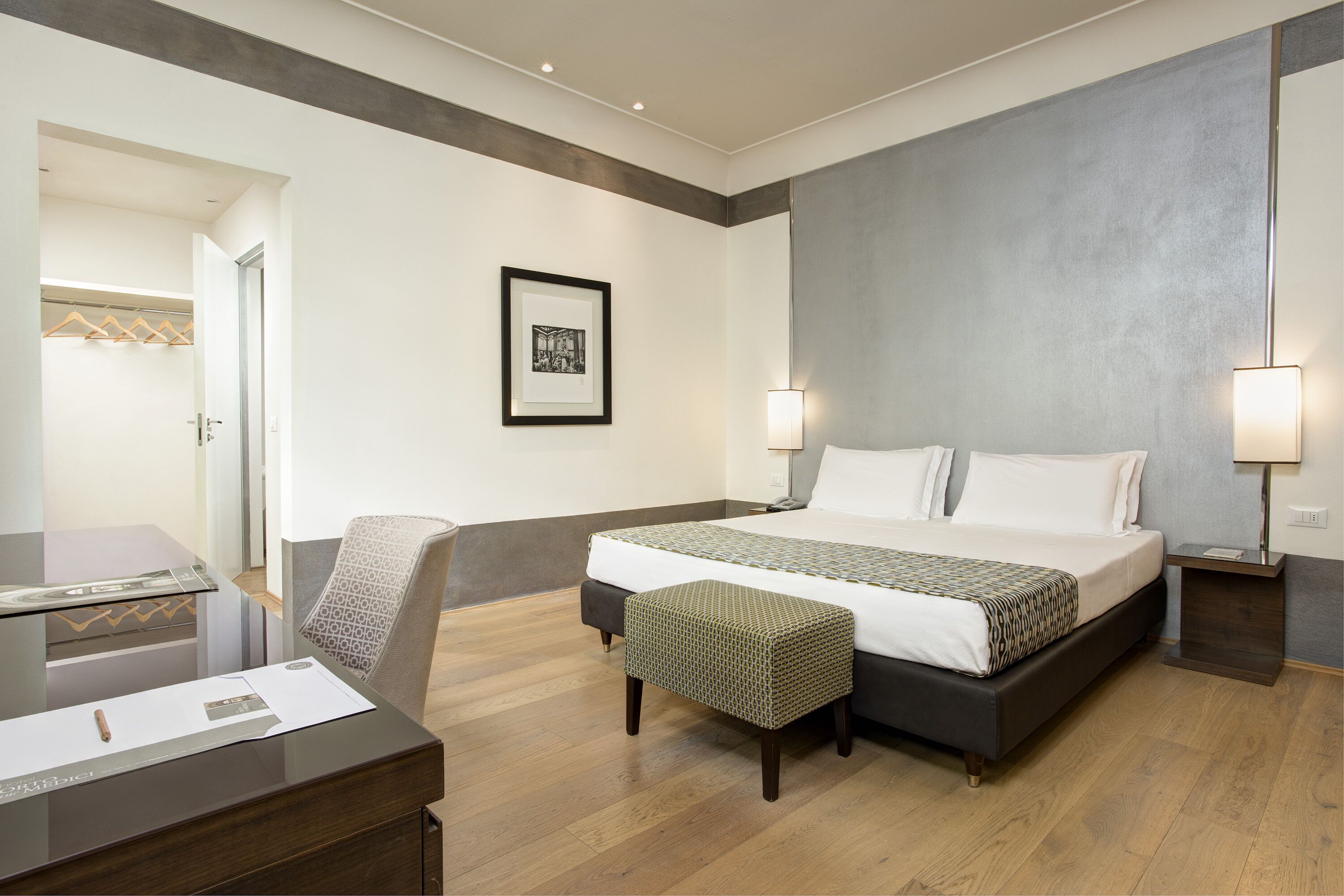 A hotel room with a bed, bedside tables, a desk, a chair, and a framed picture on the wall.