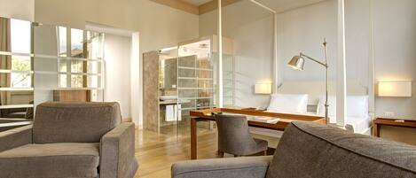A modern hotel room with a sofa, a desk, a chair, a bedside table, a lamp, and a large window.