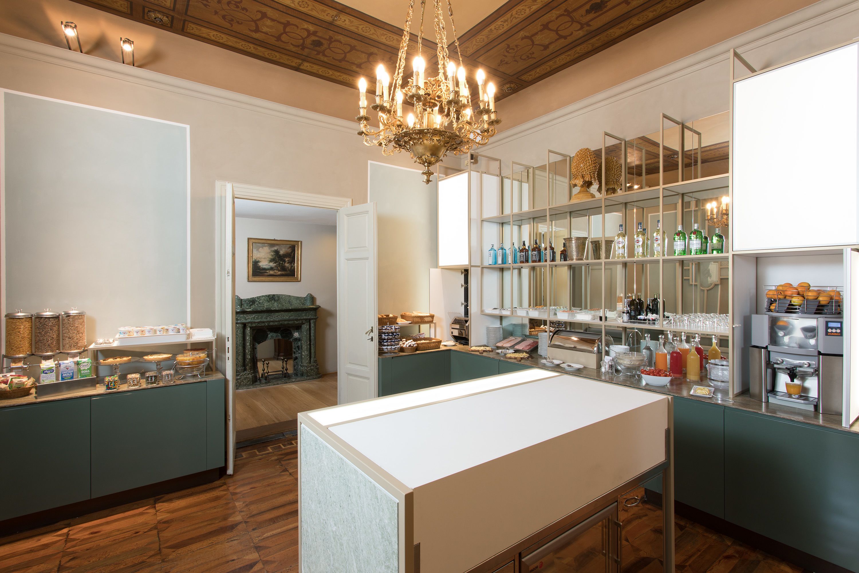 A spacious kitchen with a central island, wooden flooring, and a chandelier. There are shelves with bottles and a refrigerator with a built-in oven.