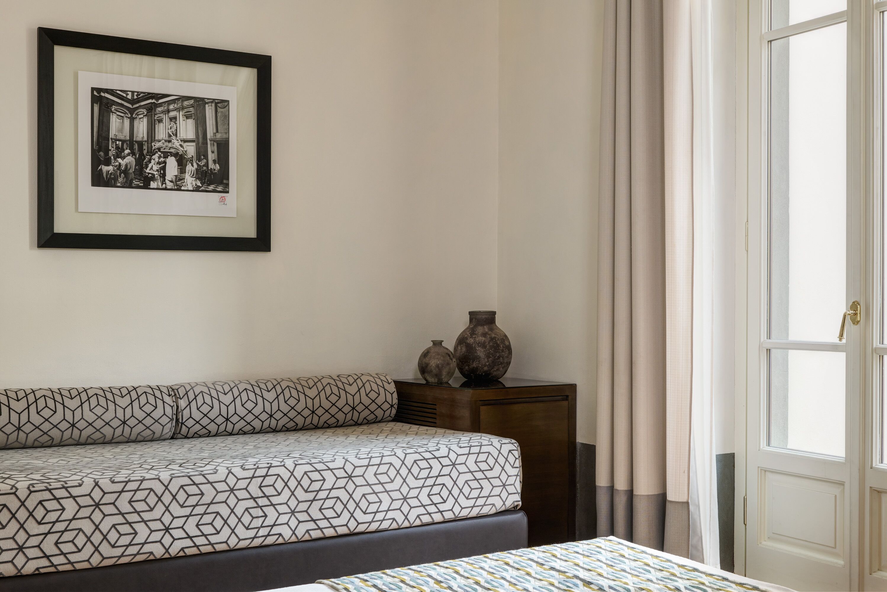 A bedroom with a bed, a framed picture on the wall, a nightstand with vases, and a door with curtains.