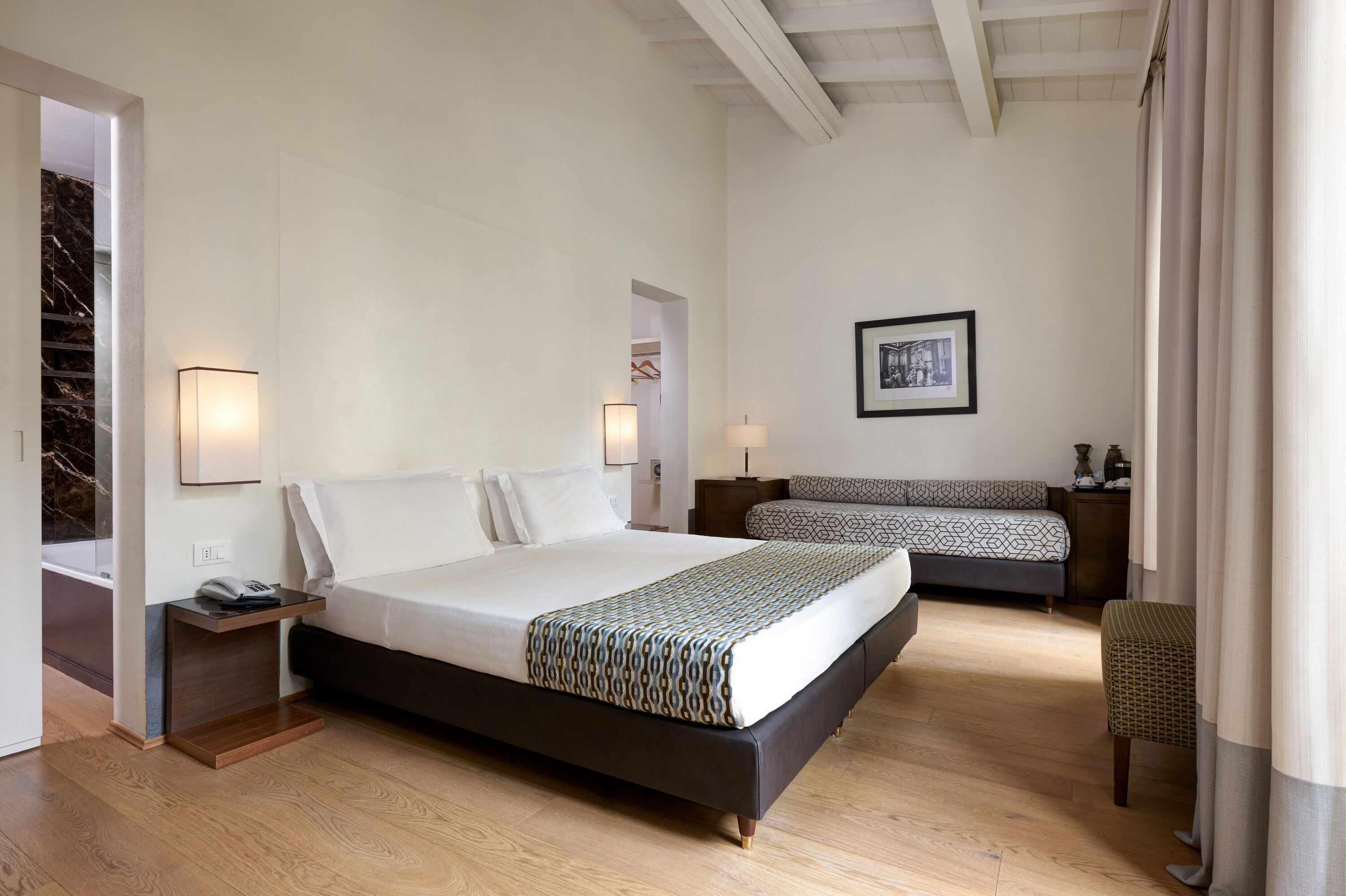 A modern hotel room with a large bed, a smaller sofa, a bedside table with a lamp, and a framed picture on the wall.