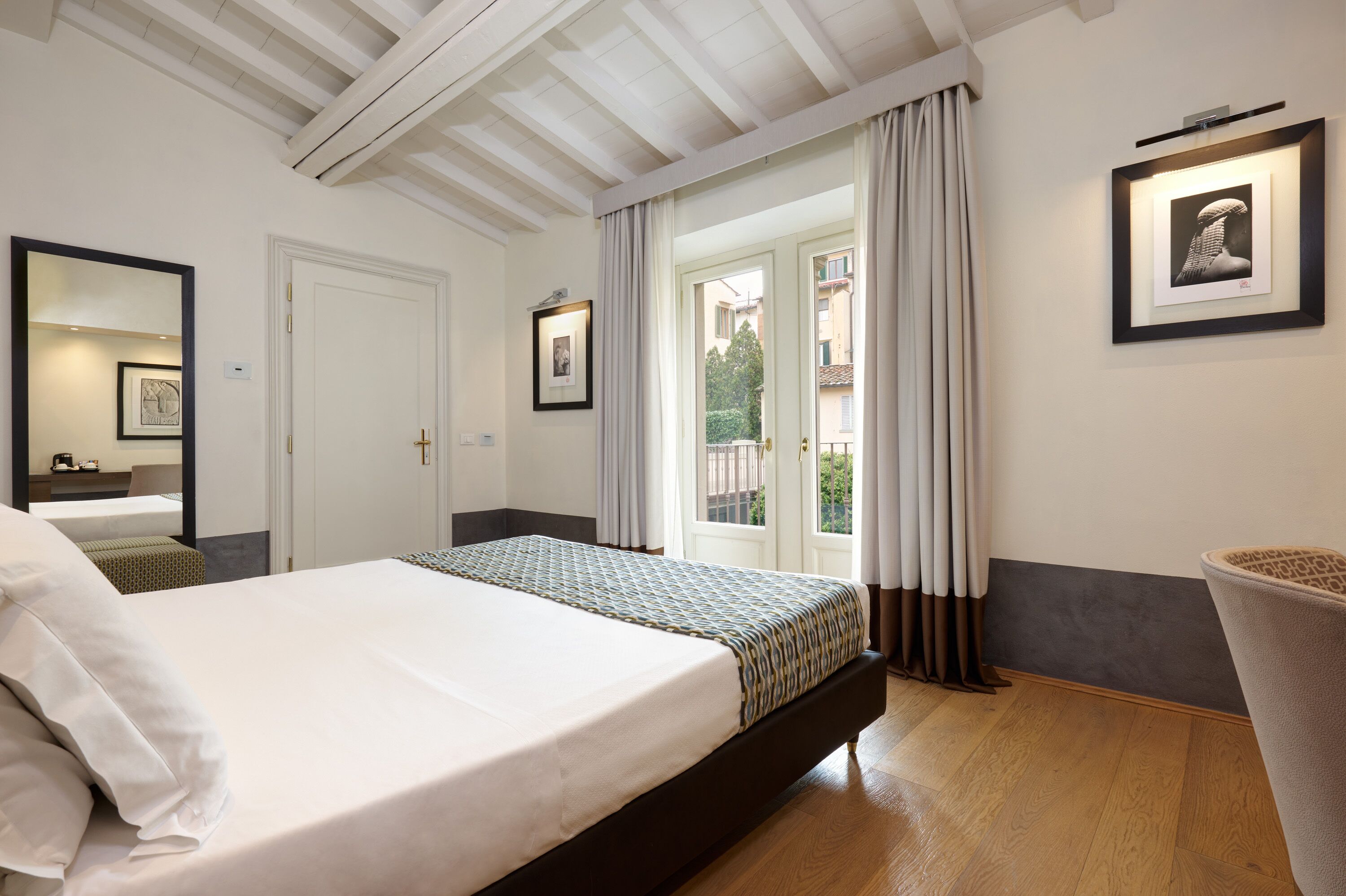 Ein Hotelzimmer mit einem großen Bett, einem Spiegel, einem gerahmten Bild und einem Fenster mit Vorhängen.