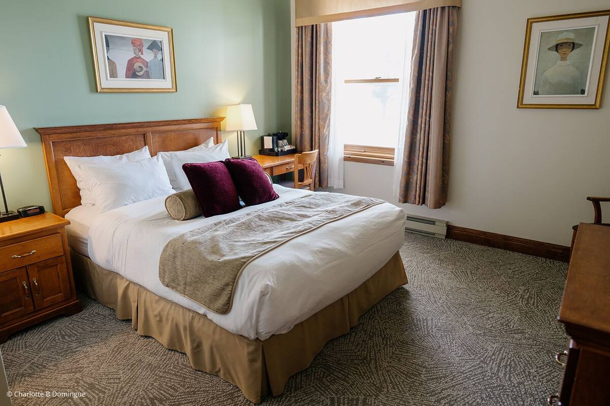 Ein Hotelzimmer mit einem großen Bett, zwei Nachttischen, einem Schreibtisch und einem Stuhl. An den Wänden hängen zwei gerahmte Kunstwerke und ein Fenster mit Vorhängen.