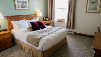Habitación de hotel con una cama grande, dos mesitas de noche, un escritorio y una silla. Se observan dos cuadros enmarcados en las paredes y una ventana con cortinas.