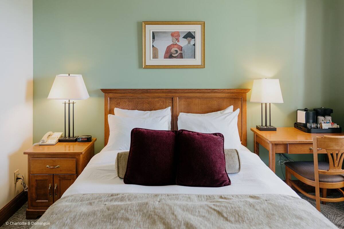 Ein Hotelzimmer mit einem Holzkopfteil, zwei Nachttischen mit Lampen, einem Schreibtisch mit Stuhl und einem gerahmten Bild an der Wand.