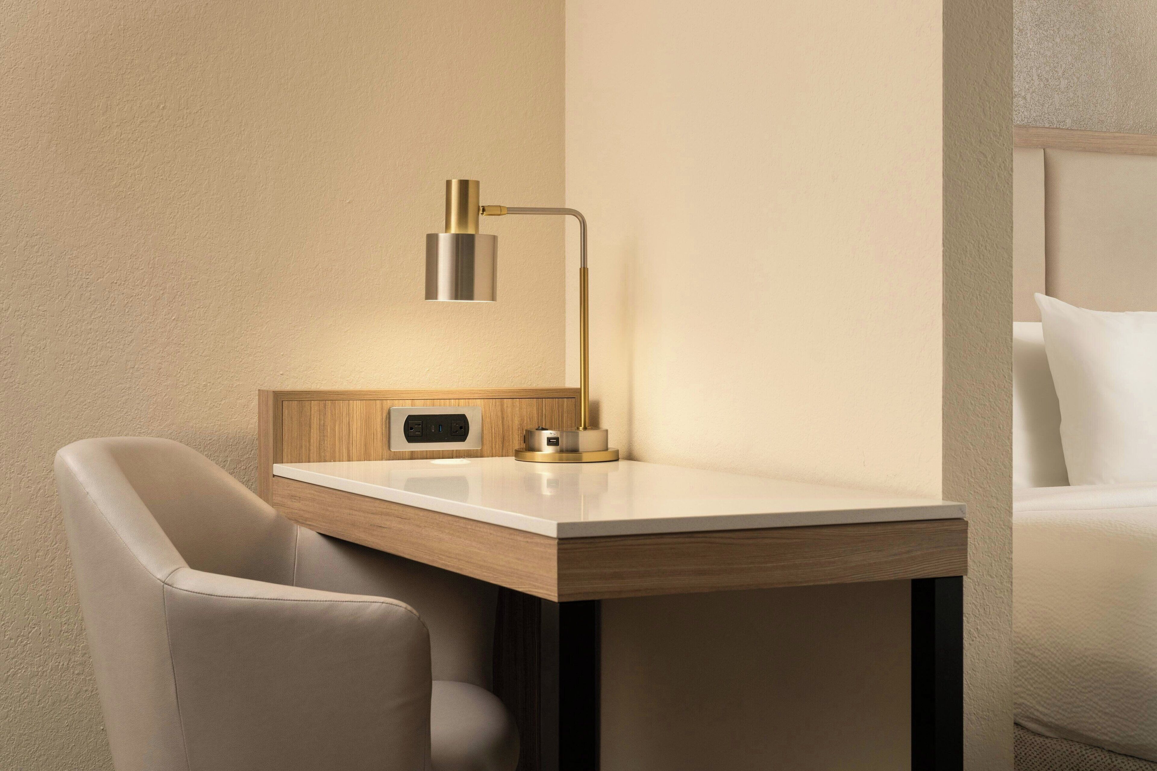 A modern hotel room with a wooden desk, a lamp, and a bed with white bedding.