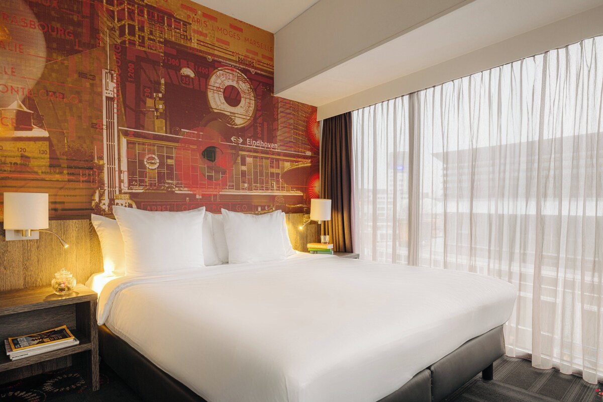 Habitación de hotel con una cama grande, una mesita de noche con lámpara, un reloj de pared y una ventana con cortinas traslúcidas.