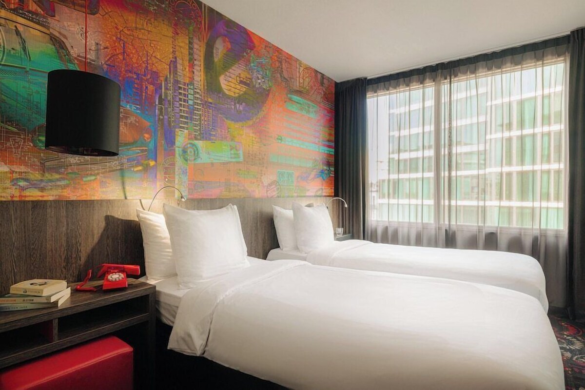 A hotel room with two beds, a nightstand with a telephone, and a colorful abstract mural on the wall.