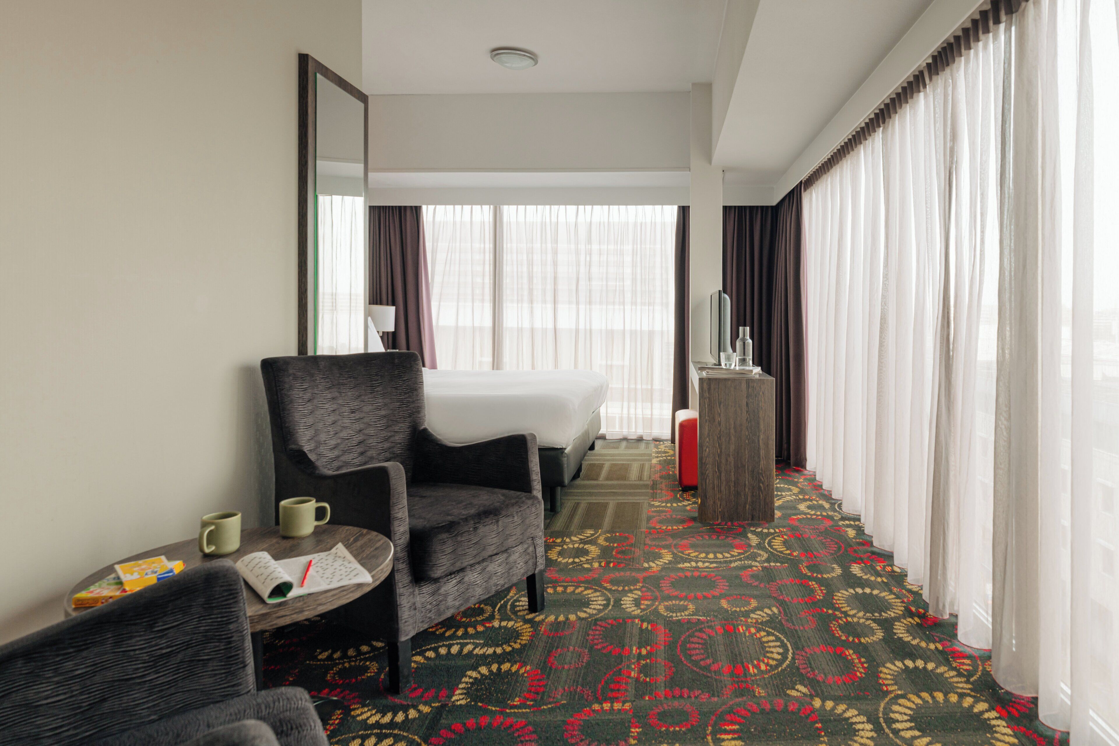 A hotel room with a bed, a chair, a small table with a book and a mug, a red ottoman, and a patterned carpet.