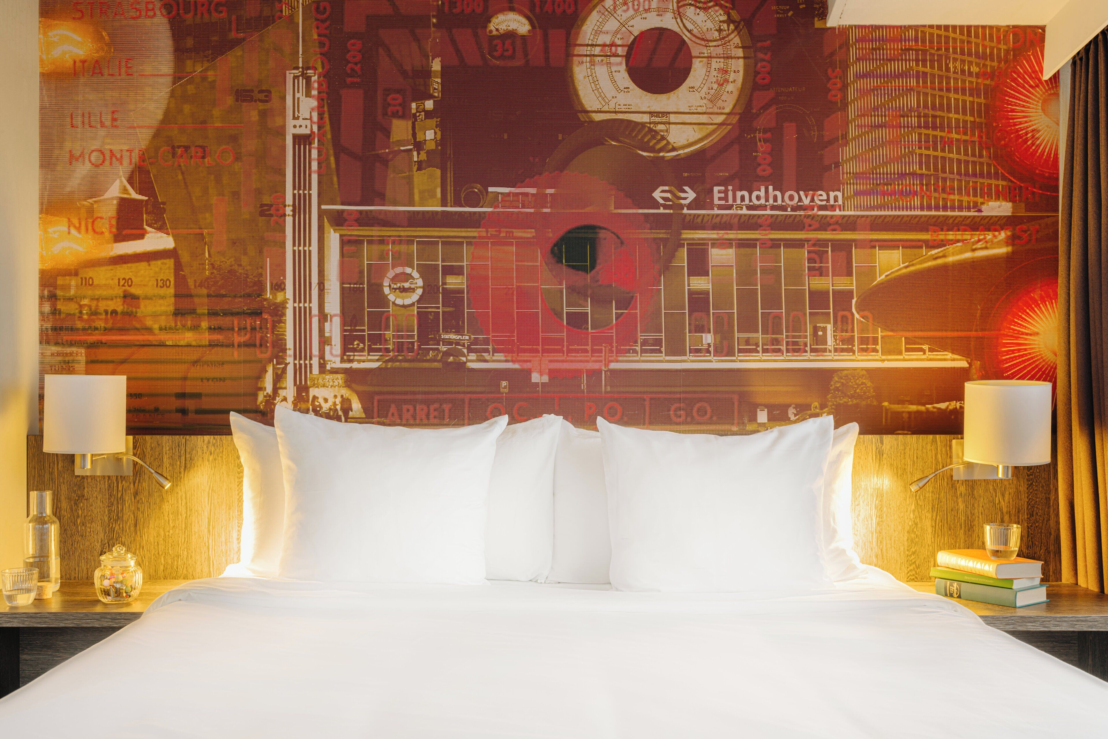 Een hotelkamer met een bed, twee nachtkastjes, een lamp en een muur met een stadsgezicht.