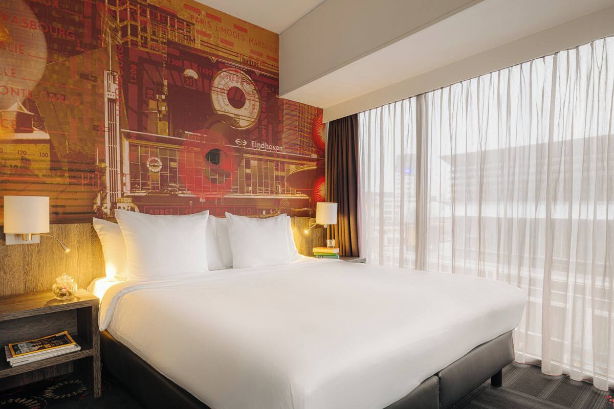 Une chambre d’hôtel avec un grand lit, une table de chevet avec une lampe, une horloge murale et une fenêtre avec des rideaux légers.