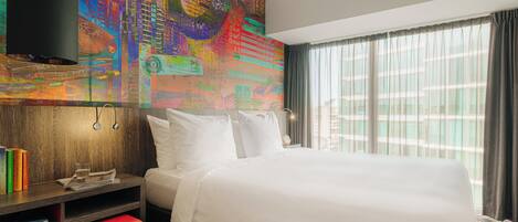 A hotel room with a large bed, a wooden nightstand, a red bench, a colorful abstract wall art, and a window with curtains.