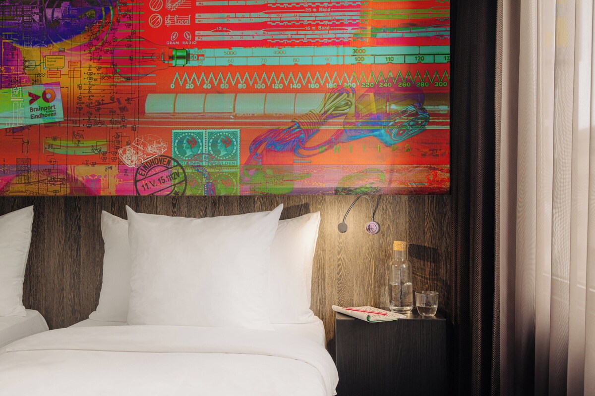 Habitación de hotel con cama, mesita de noche y pared decorada con arte colorido.