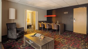 A hotel room with a gray armchair, a wooden coffee table with a book and a glass, a bar area with chairs, and a patterned carpet.