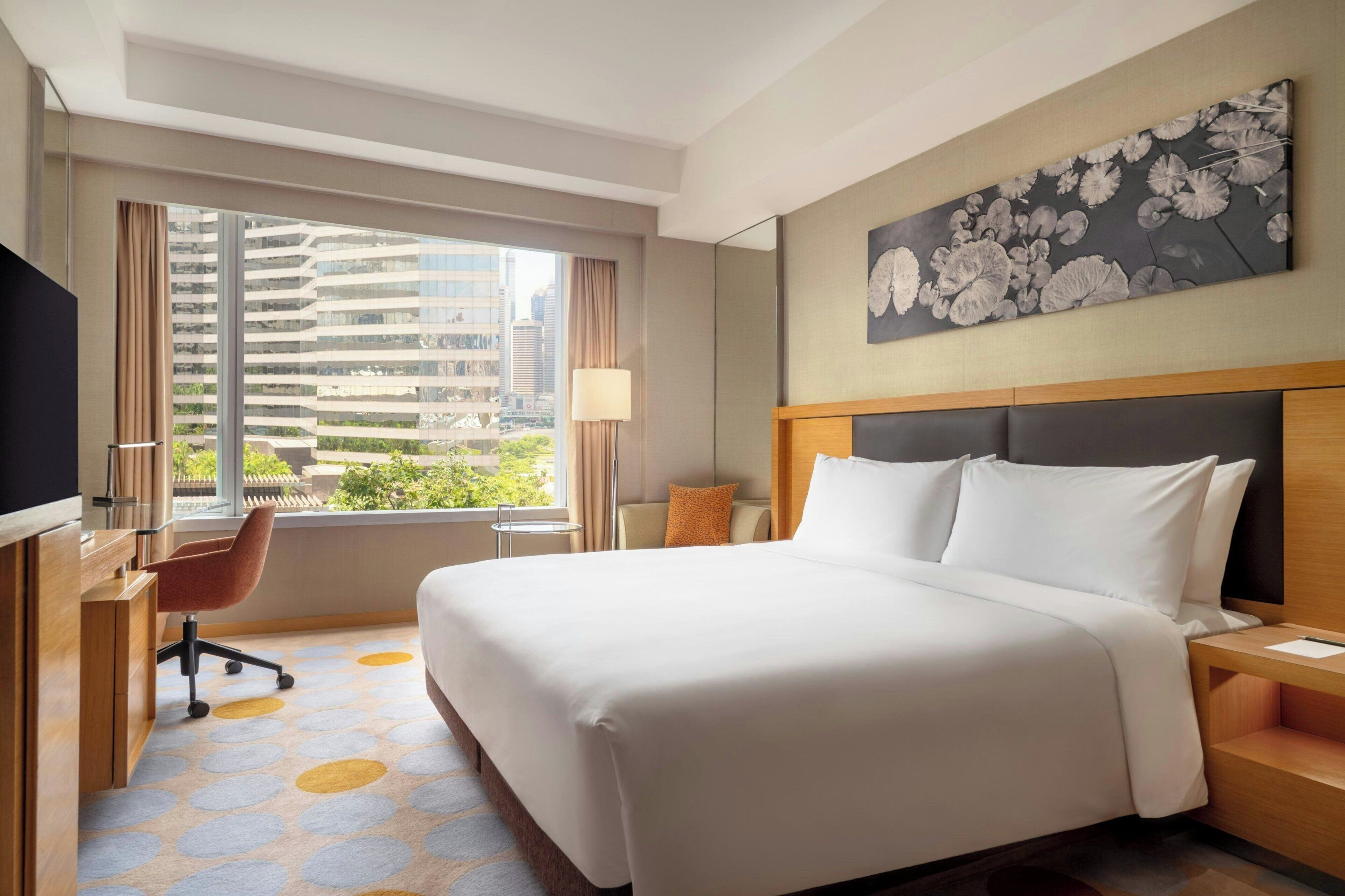 A hotel room with a large bed, a desk, a chair, a TV, and a window with a city view.