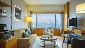 A modern hotel room with a city view, a sofa, a round table, a TV, and a desk with a lamp.