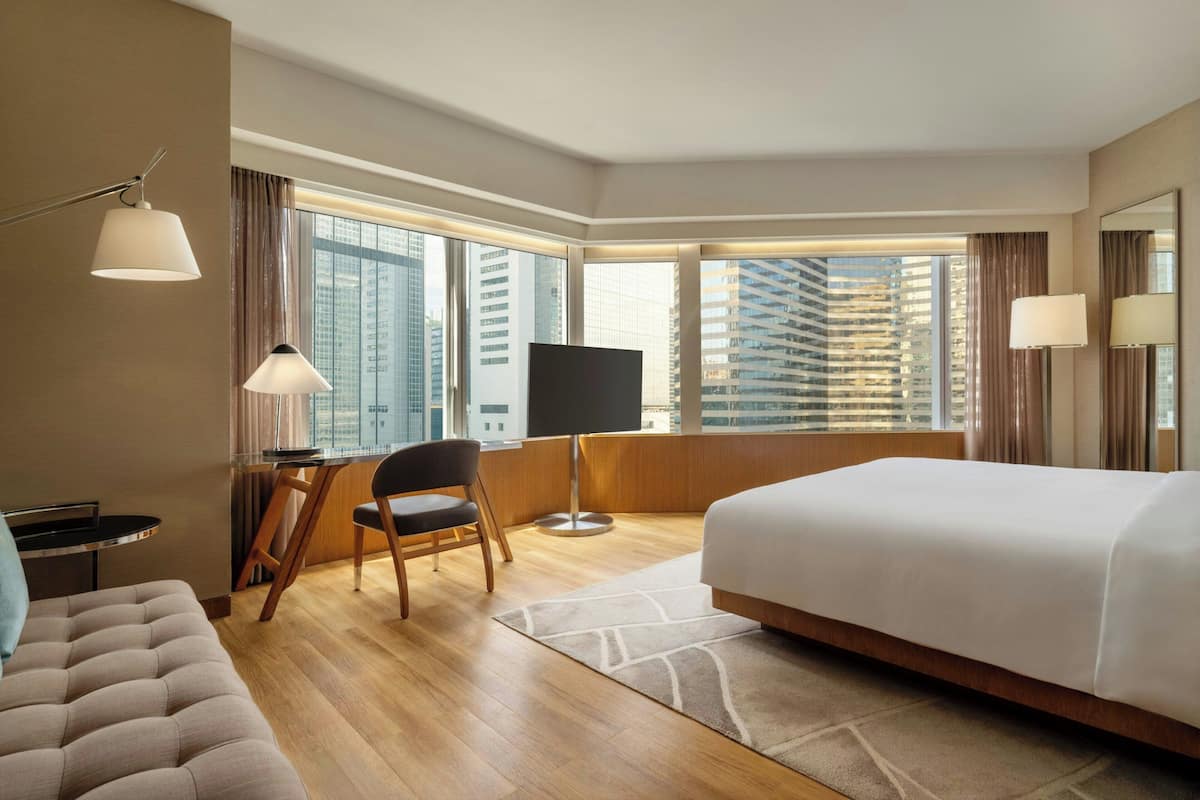 A hotel room with a large bed, a desk with a chair, a TV, and a view of tall buildings through large windows.