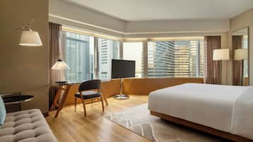 Ein Hotelzimmer mit einem großen Bett, einem Schreibtisch mit Stuhl, einem Fernseher und Blick auf hohe Gebäude durch große Fenster.