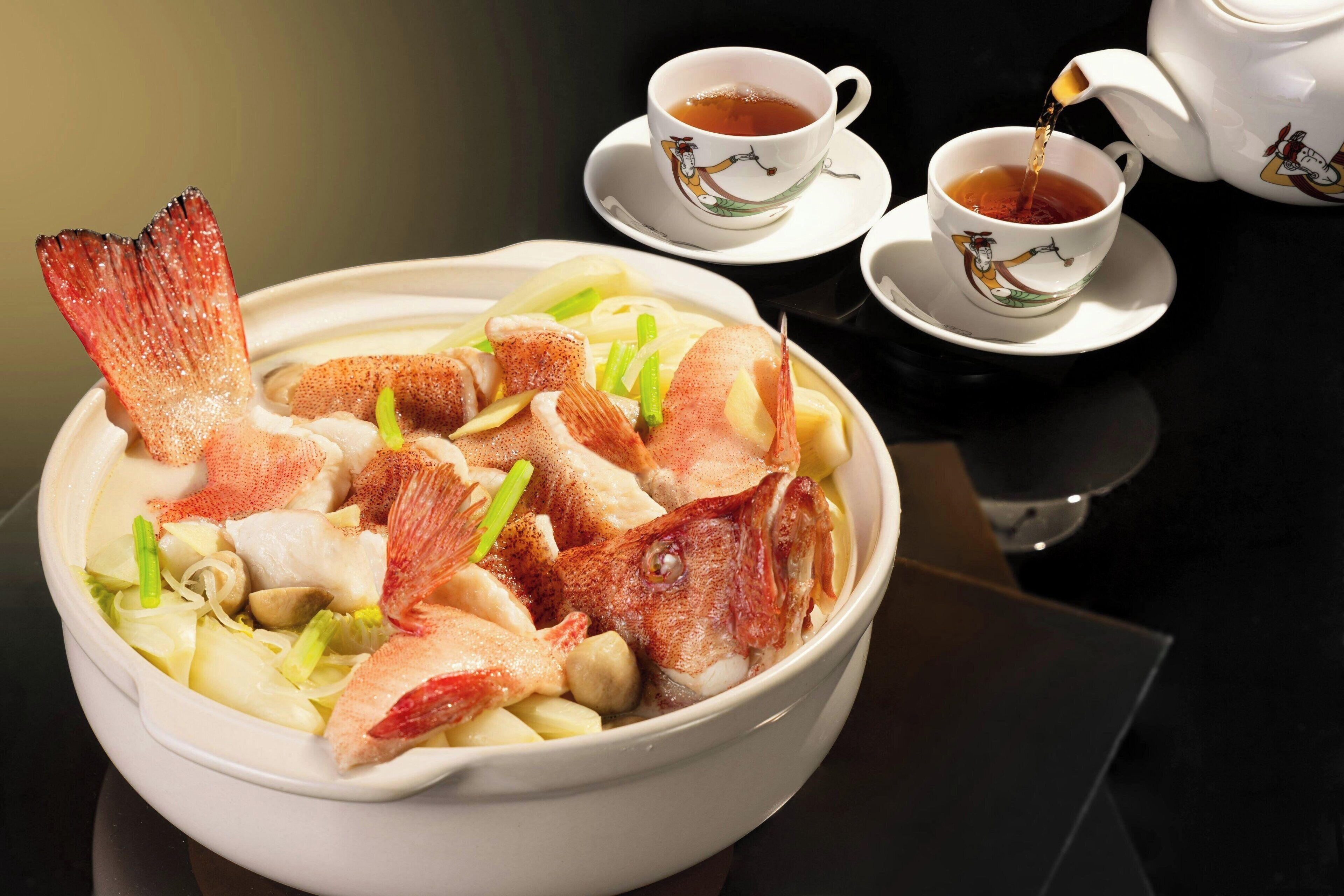 A bowl of soup with fish, vegetables, and a cup of tea on a table.